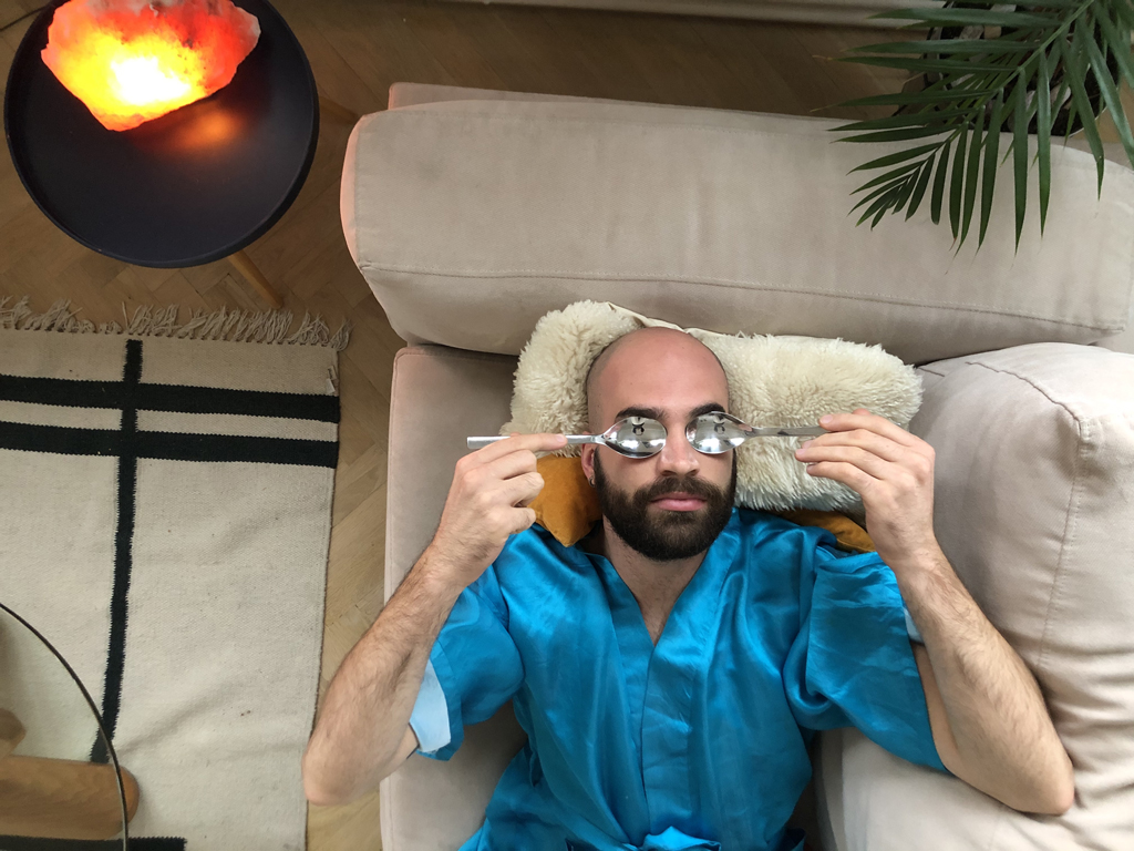 A image of a man lying on a couch holding metal spoons over his eyes